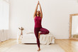 © Anatoliy Karlyuk - Full length portrait of beautiful young Afro American woman with fit healthy body raising hands and placing sole of foot on her thigh, doing tree yoga stance or vriksasana in cozy bedroom interior