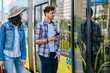 © Iryna - Lifestyle photo of young woman and man passengers holds phones waiting while opening door to tram. Portrait of female traveler with backpack talking with handsome guy on tram stop.