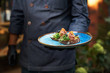canvas print picture - delicious raw beef tartare on bruschette, the dish is held in hand by a cook in a blue uniform.