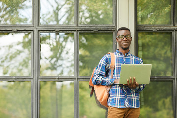 Waist up portrait of smart African student holding laptop posing outdoors in college campus and looking at camera, copy space