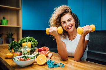 Wall Mural - Portrait of fit and strong girl, model and fitness trainer holding dumbbells, with fresh salade on table, healthy eat time.