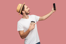 Side View Portrait Of Handsome Bearded Young Hipster Man In White Shirt And Casual Hat Posing And Making Selfie Photo With Smile, Blogger Lifestyle. Indoor,isolated, Studio Shot, Pink Background