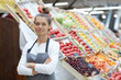 Waist up portrait of pretty young saleswoman looking at camera while posing by fresh fruits and vegetables at supermarket, copy space