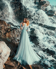 sweet lady in light long blue flying dress, water nymph stands near large waterfall in spray of clean cold aqua. mermaid with blond dyed hair stands with her back to camera and turned face