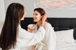 © Anton - Two young beautiful women with dark hair in white bathrobes sitting on bed happily making cosmetic mask together in modern hotel