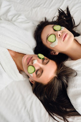 Wall Mural - Two young beautiful women with dark hair in white bathrobes with cosmetic mask and slices of cucumber on faces dreamily lying in bed in modern cozy hotel