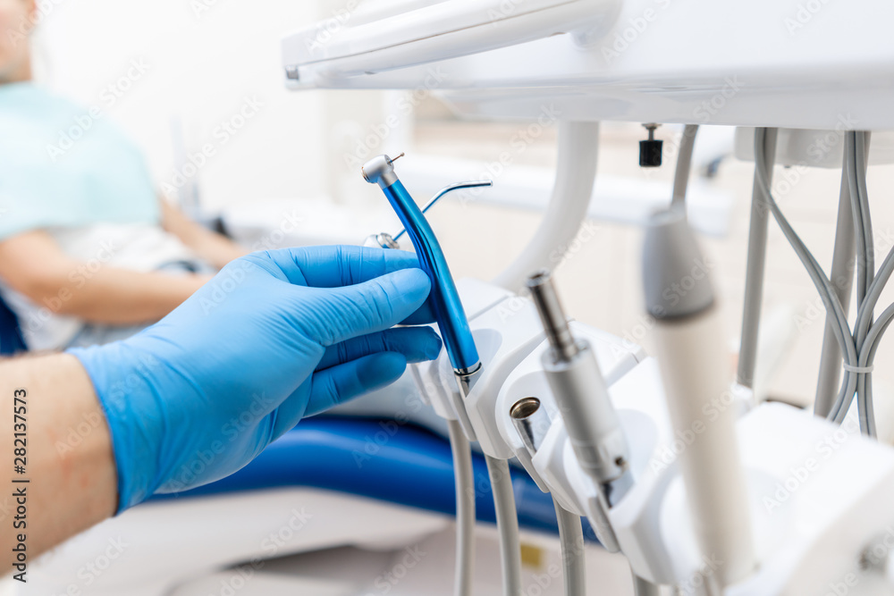 Close-up hand of dentist in the glove holds dental high speed turbine. The patient in blue chair at the background. Office where dentist conducts inspection and concludes. - obrazy, fototapety, plakaty 