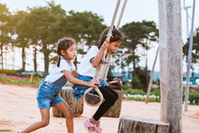Happy Asian Child Girl Having Fun To Play On Wooden Swings With Her Sister In Playground With Beautiful Nature