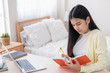 © weedezign - Asian female freelancer reading and jot down note on book while working with laptop on table in bedroom at home.Work at home concept.work from home concept.