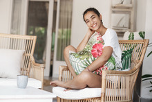 Happiness, Vacation And Beauty Concept. Cheerful Tender Young Woman In White T-shirt Sitting Legs Crossed On Rattan Armchair, Hugging Pillow, Laughing Cute, Enjoying Summertime Luxury Resort Trip