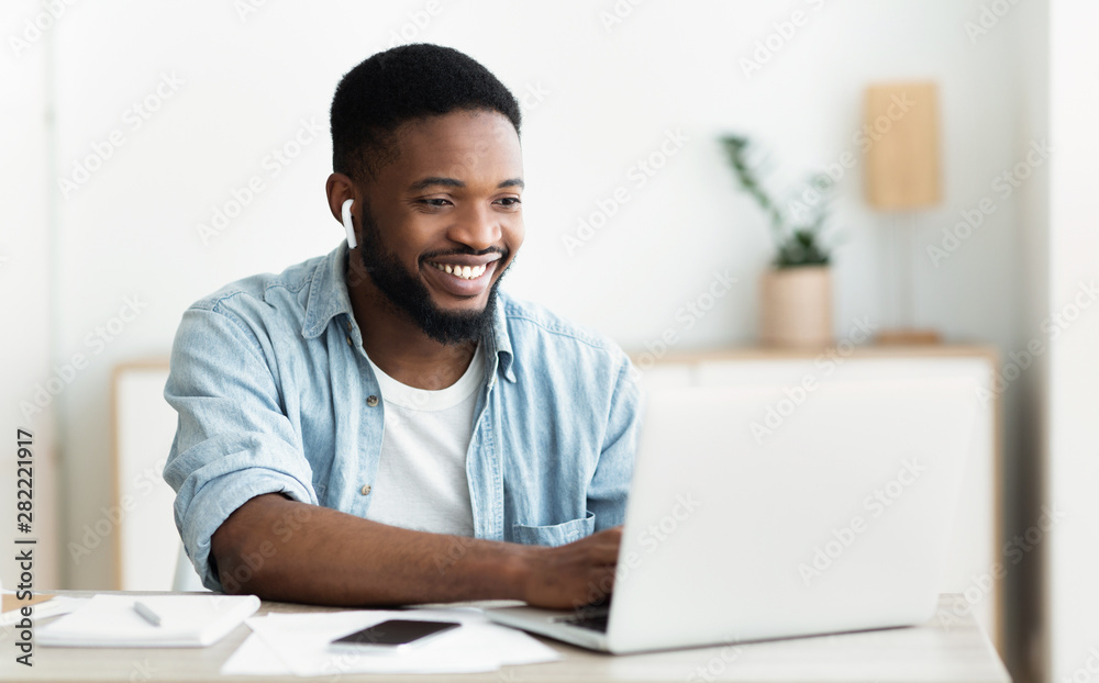 Smiling african-american guy in earphones studying foreign language online - obrazy, fototapety, plakaty 