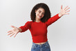 © Cookie Studio - Girl wanna hold you. Charming pleasant lovely curly-haired woman wearing red stylish cropped top spread hands hug cuddles smiling friendly cute inviting embraces, standing white background