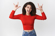 © Cookie Studio - Rocking roll baby. Excited funny emotive happy smiling young woman curly dark hair attend awesome rock concert party show heavy metal sign stick tongue thrilled fool around, rejoicing