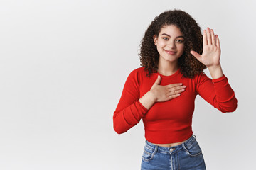 Wall Mural - Promise tell truth. Charming happy relaxed sincere young curly-haired girl pleadging oath swear hold hand heart raise palm making statement smiling happily, standing white background