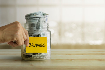 Money savings concept. Man is holding a sticky note with the word “savings” stick on glass jar full of dollar banknotes.