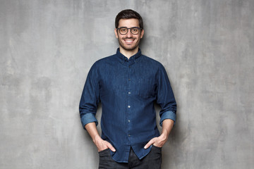 Wall Mural - Young smiling man wearing trendy glasses and denim shirt, leaning on gray textured wall