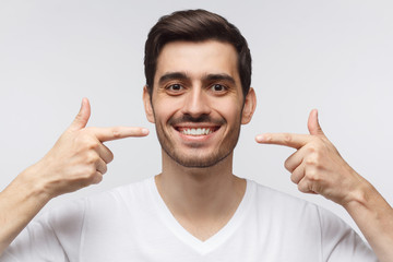 Portrait of smiling young man pointing to his perfect natural white teeth after whitening treatment. Dental clinic ad concept