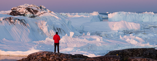 Wall Mural - Travel wanderlust adventure in Arctic landscape nature with icebergs - tourist person looking at view of Greenland icefjord - aerial photo. Man by ice and iceberg, Ilulissat Icefjord.