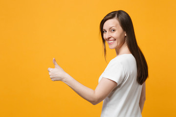 Portrait of smiling young woman in white casual clothes looking back and showing thumbs up isolated on bright yellow orange wall background in studio. People lifestyle concept. Mock up copy space.