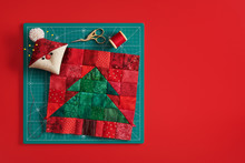 Christmas Tree Patchwork Block, Pincushion Like Santa, Scissors, Spool Of Thread On Craft Mat, Red Background