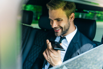 Close up of handsome young man in full suit talking on speakerphone of smart phone and smiling while sitting in the car