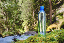 Plastic Bottle Blue Cap With Fresh Drinking Water On A Background Of Green Forest Stands On A Stone With Moss. The Concept Of Pure Natural Mountain Water