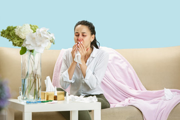 Wall Mural - Young woman suffering from hausehold dust or seasonal allergy. Sneezing in the napkin and sitting surrounded by used napkins on the floor and sofa. Taking medicines with no result. Healthcare concept.