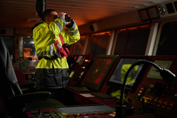 Navigator. pilot, captain as pat of ship crew performing daily duties with VHF radio, binoculars on board of modern ship with high quality navigation equipment on the bridge on sunrise.