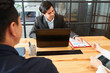 © DragonImages - Young male leader sitting at the table and working on laptop computer and holding a business meeting for his employees at office
