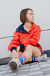 A short haired woman in coral windbreaker and colorful socks sitting on road near grey wall