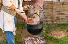 Over A Fire On The Hook Hangs A Large Cauldron. Cook With A Ladle Pouring The Stew In A Bowl Made From A Large Loaf Of Bread