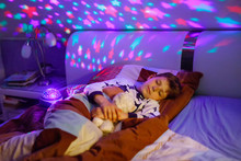Little Kid Boy Sleeping In Bed With Colorful Lamp. School Child Dreaming And Holding Plush Toy. Kid Angry Of Darkness. Night Light Changing Colors, Stars And Moon And Playing Music.