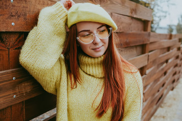 Pretty young red hair ginger hair  sweater and yellow beret hat posing in park near wooden wall. Beautiful woman wearing trendy seasonal clothes in autumn.