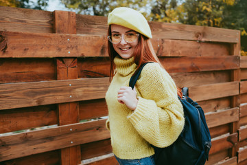 Pretty young red hair ginger hair  sweater and yellow beret hat posing in park near wooden wall. Beautiful woman wearing trendy seasonal clothes in autumn.