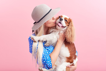 Wall Mural - Portrait of smiling young blond woman in summer hat embracing king Charles spaniel dog. owner and pet relations concept. Veterinary health. Isolated front view on pink background.