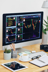 computer screen with stock market graphs, cup of green tea, tablet computer and planner of desk fina
