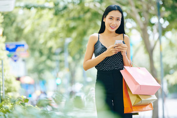 Poster - Portrait of Asian brunette in beautiful suit with shopping bags using mobile phone while walking along the street and smiling at camera