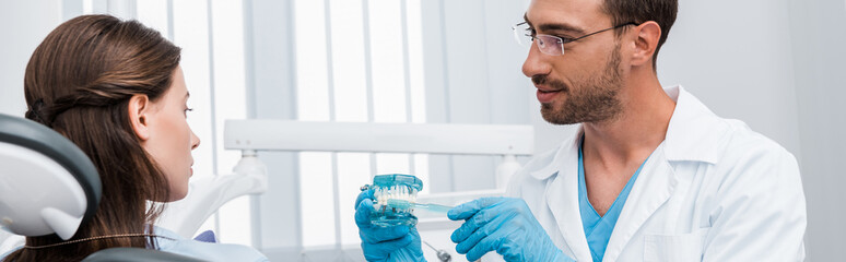 Wall Mural - panoramic shot of dentist in latex gloves holding toothbrush near teeth model and attractive girl