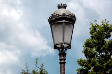 Black Iron Street Lamp In Retro Style With A Glass Shade In The Background In The Blur Sky With Clouds And Tree Branches.