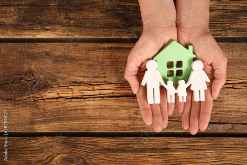 Fotomural Woman holding figures of family and green house in hands on wooden background, top view