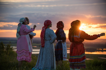 Wall Mural - People in traditional russian clothes standing on the field and looking at the bright sunset