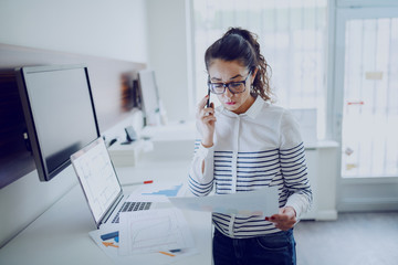 Serious Caucasian businesswoman dressed casual talking on the phone and looking at paperwork while standing at office.