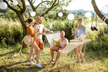 Wall Mural - Young friends relaxing with drinks, sitting on the hammmock on the backyard or garden during a festive meeting on the sunset