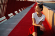 © vitaliymateha - Bright summer lifestyle portrait of young pretty woman in eyewear, red skirt and white T-shirt, sitting on a bright red floor on the bridge.