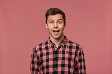 Wall Mural - Portrait of young attractive happy amazed man in checkered shirt, looks at the camera with surprised expression, stands over pink background with wide open mouth.