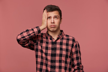 Poster - Portrait of young attractive man in checkered shirt, looks up, stands over pink background and touches head, looks tired and sad.