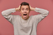 Close up of young scared man in blank long sleeve, stands over pink background with closed eyes and screaming, looks afraid and unhappy, touches head.