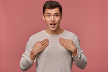 Wall Mural - Portrait of young attractive surprised man wears in blank t-shirt, looks at the camera with happy expression, point at himself, isolated over pink background.