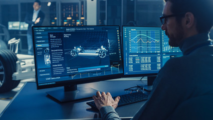 Fototapete - Professional Engineer Works on a Computer with a 3D CAD Software and Tests the Electric Car Chassis Prototype with Wheels, Batteries and Engine Standing in a High Tech Development Laboratory.
