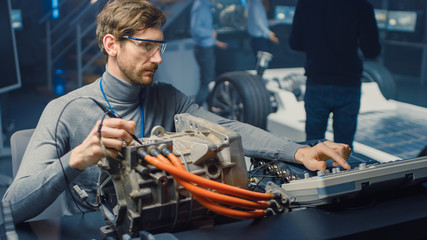 Fototapete - Professional Automotive Engineer in Glasses with a Computer and Inspection Tools is Testing an Used Electric Engine in a High Tech Laboratory with a Concept Car Chassis.